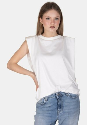 Giovane donna con capelli lunghi indossa una canotta bianca senza maniche con spalle imbottite e jeans azzurri, in piedi con una mano sul fianco.