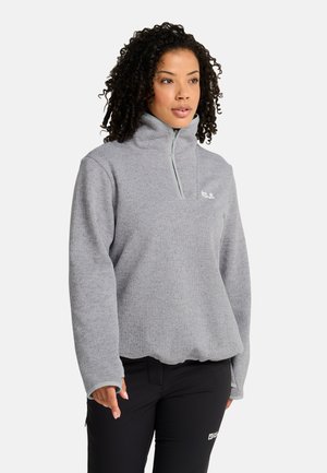 Frau mit lockigem Haar, die eine hellgraue Fleecejacke mit Viertelreißverschluss und schwarze Hose trägt, steht vor einem schlichten hellen Hintergrund.