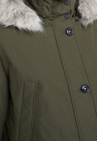 Olivově zelená parka s kapucí s umělým kožíškem, opatřená černými knoflíkovými zapínáními, předním kapsou a texturovaným povrchem látky.