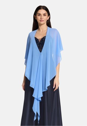 Vrouw die een doorschijnende lichtblauwe sjaal draagt over een zwarte jurk met pailletten, staand tegen een effen witte achtergrond.