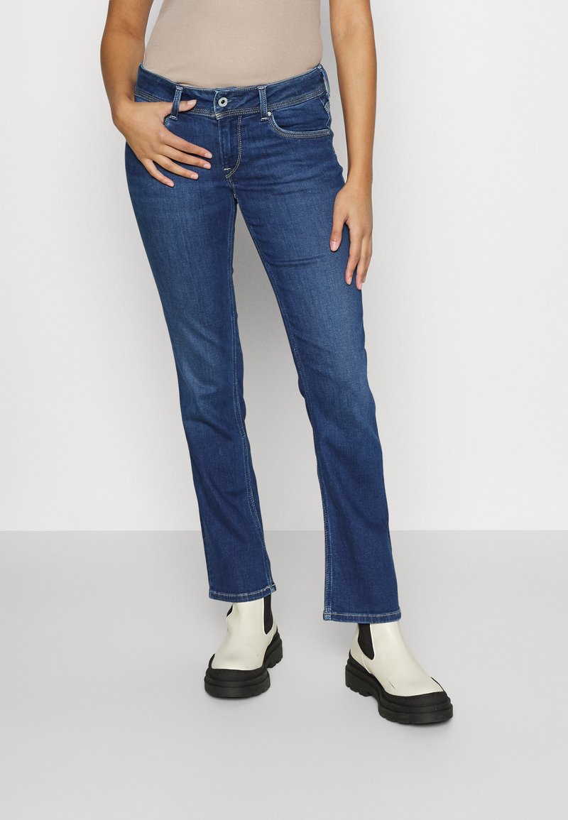 Jean en denim bleu avec une coupe droite, présentant un style classique à cinq poches et une fermeture à bouton ; porté avec des bottines noires et blanches.
