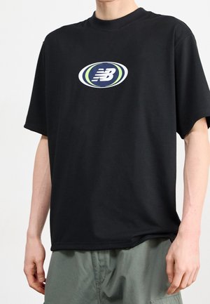 Mężczyzna w czarnej, oversize'owej koszulce z logo składającym się z białego napisu "NB" wewnątrz granatowo-zielonego owalu, w połączeniu z szaro-zielonymi spodniami.