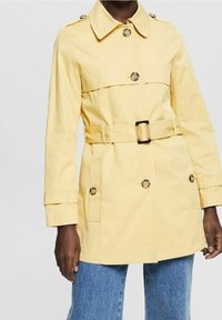 Trench-coat jaune avec une taille ceinturée, de gros boutons, un col et deux poches latérales. Confectionné en matériau léger avec des manches longues.