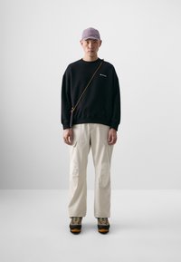 Sweatshirt noir avec un petit logo, pantalons crème avec poches avant, et chaussures de randonnée gris-orange. Casquette violet clair et sac en bandoulière.