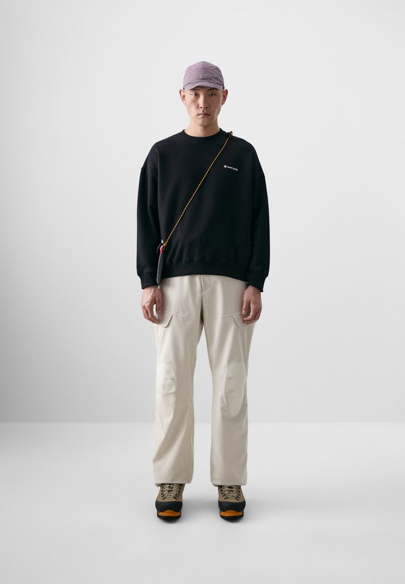 Sweatshirt noir avec un petit logo, pantalons crème avec poches avant, et chaussures de randonnée gris-orange. Casquette violet clair et sac en bandoulière.