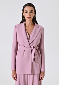 Helles rosa Blazer mit langen Ärmeln, einem V-Ausschnitt, Wickeldesign, Taillengürtel und silbernen Hardware-Details. Glatte Textur mit taillierter Passform.