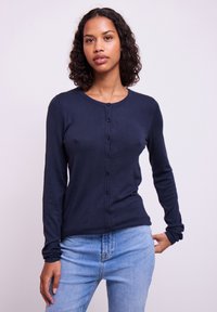 Marineblauer Langarm-Cardigan aus Strickstoff, mit durchgehender Knopfleiste, rundem Ausschnitt, figurbetontem Schnitt und gerippten Bündchen, getragen über hellblauen Jeans.