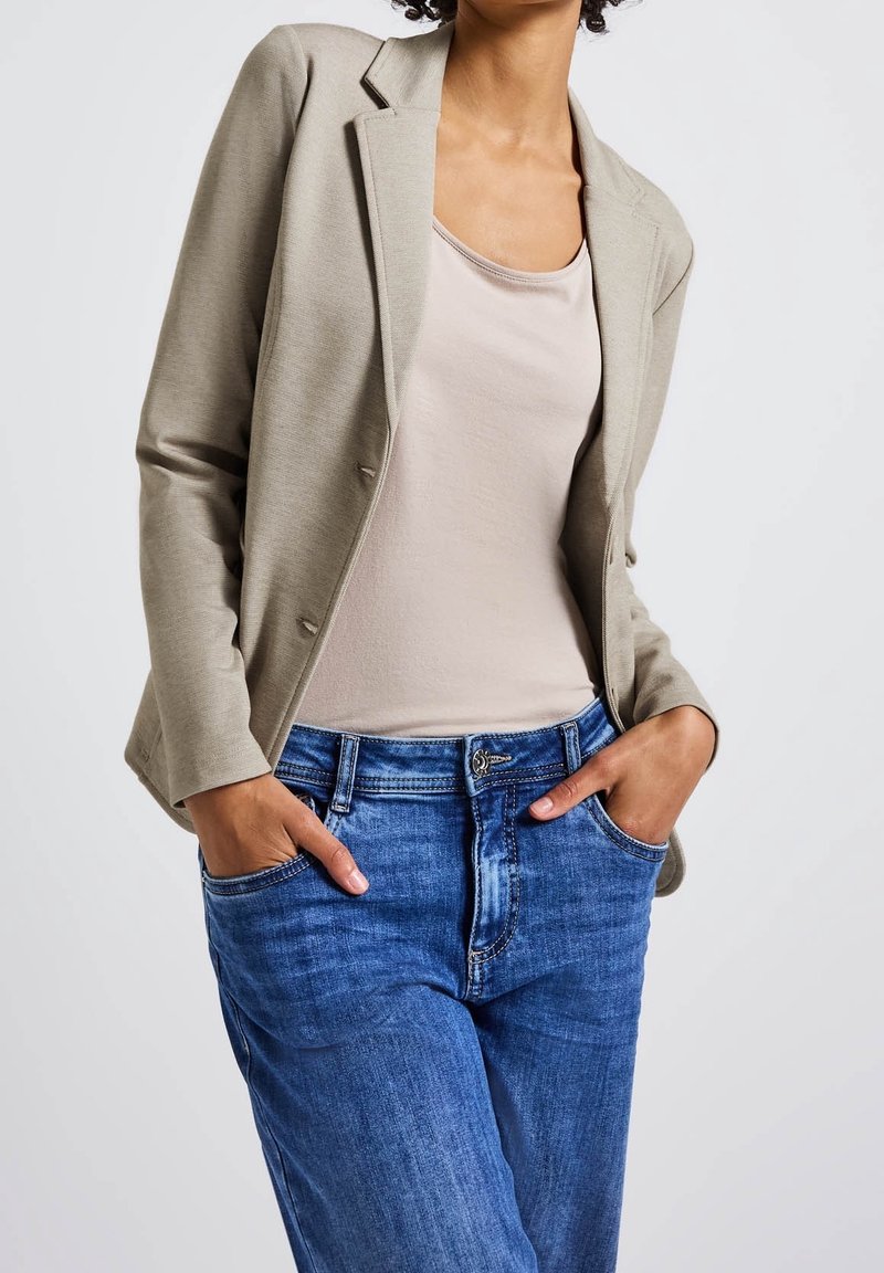 Femme les mains dans les poches, portant un blazer beige, un débardeur rose clair et un jean bleu sur fond blanc uni.
