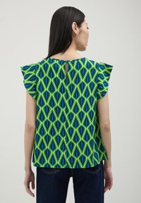 Blusa verde con motivo a goccia blu e nera, maniche corte a volant e dettaglio a chiave sulla schiena. Indossata con jeans scuri.