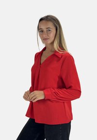 Blusa rossa a maniche lunghe con scollatura a V e colletto, caratterizzata da dettagli plissettati sulle maniche e una vestibilità ampia. Realizzata in tessuto testurizzato.