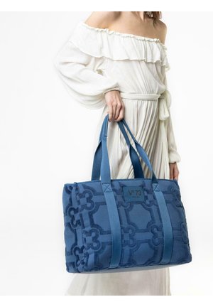 Mujer con vestido blanco de hombros descubiertos sosteniendo un gran bolso tote azul con estampado, con asas dobles y etiqueta de la marca.