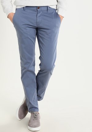 Lichtblauwe katoenen chinos met een rechte pijp, voorzien van een bruine knoopsluiting en zijzakken. Gecombineerd met grijze sneakers.