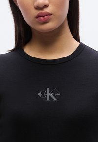 Μαύρο ριμπ φούτερ με στρογγυλή λαιμόκοψη, με ασημί λογότυπο που γράφει "Calvin Klein" και στιλιζαρισμένο "ck" στην π εριοχή του στήθους.