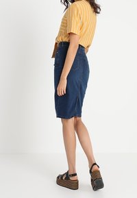 Falda lápiz de mezclilla con un corte a medio largo, que presenta una abertura trasera para facilitar el movimiento. Combinada con una blusa amarilla a rayas y sandalias de cuña negras.