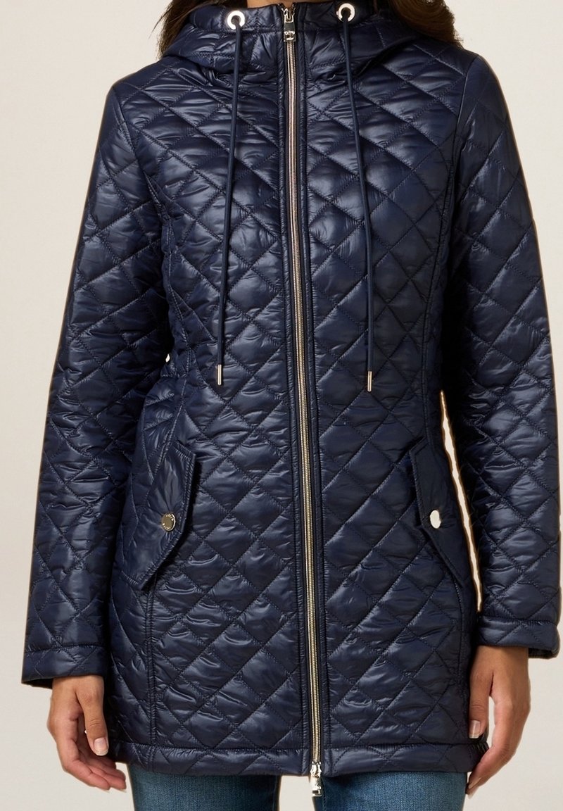 Abrigo acolchado azul marino con cierre de cremallera, capucha con cordón, dos bolsillos laterales y acentos metálicos. Presenta una textura suave y brillante.