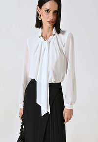 Blusa bianca con maniche lunghe e dettaglio a nodo sul davanti, realizzata in tessuto trasparente, abbinata a una gonna nera plissettata. Accenti dorati sullo scollo.