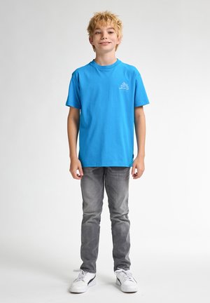 Blå bomulds t-shirt med korte ærmer og rund hals, med et lille grafikdesign på brystet. Grå jeans og hvide sneakers.