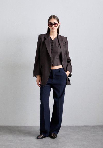Dunkelbrauner Doppelreiher-Blazer, kurzes schwarzes Top mit Knöpfen, marineblaue weit geschnittene Hose und schwarze Schuhe mit Gittermuster. Große dunkle Sonnenbrille.