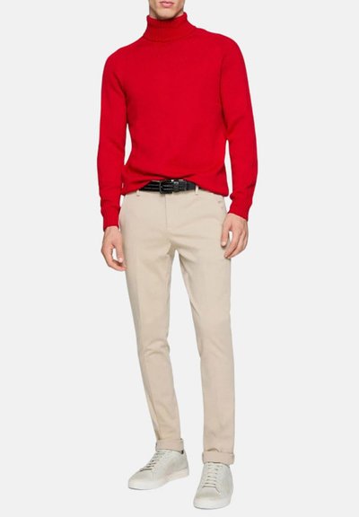 Maglione a collo alto rosso lavorato a maglia, pantaloni beige e sneakers bianchi. Texture liscia, design aderente, con un accento di cintura nera.