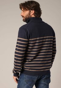 Pull rayé bleu marine et beige avec un col haut, fabriqué en tissu doux. Porté avec un jean bleu, avec des poches arrière ajustées.
