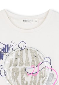 Biała koszulka z okrągłym, błyszczącym srebrnym wzorem z napisem "SHINE" w pogrubionych literach. Akcenty obejmują fioletowe i różowe elementy wokół wzoru.