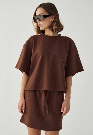 Kobieta w dużym brązowym t-shircie i pasującej spódnicy z sznurkiem, w dużych czarnych okularach przeciwsłonecznych, stojąca na jednolitym tle.