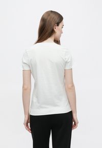 Λευκό βαμβακερό t-shirt με κοντά μανίκια, στρογγυλή λαιμόκοψη και ίσια ράχη, φορεμένο με μαύρο παντελόνι. Απλός σχεδιασμός, λεία υφή.