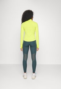 Femme portant un haut à manches longues jaune fluo et un legging vert foncé avec des panneaux en maille, se tenant de dos sur un fond clair.