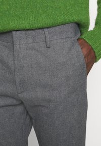 Pantalon gris sur mesure en mélange de laine texturée, avec poches avant et design à plat, associé à un pull en maille vert.