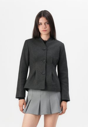 Veste ajustée gris foncé avec un col montant, quatre boutons et une taille structurée. Design ajusté avec des manches longues. Complétée par une jupe plissée grise.