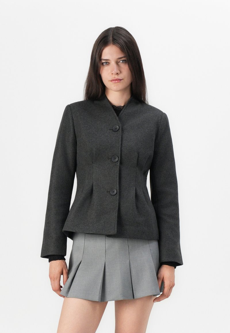Dunkelgrau maßgeschneiderte Jacke mit hohem Kragen, vier Knöpfen und strukturiertem Taillenbereich. Fitted-Design mit langen Ärmeln. Ergänzt durch einen gefältelten grauen Rock.