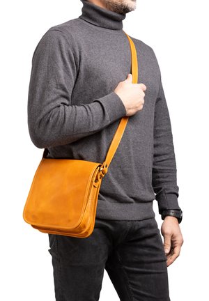 Sac en bandoulière en cuir orange avec une texture lisse, une forme structurée et une sangle ajustable, porté en travers sur un pull encolure cheminée gris.