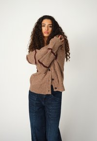 Brauner Strickcardigan mit kastanienbraunen Streifen, Knopfverschluss und lockerer Passform, kombiniert mit weit geschnittenen, dunklen Jeans. Die Textur wirkt weich und warm.