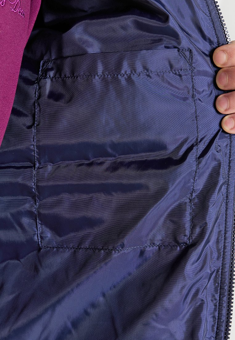 Une main tenant une veste violette ouverte montrant une poche rectangulaire cousue à l'intérieur sur la doublure lisse et brillante de couleur bleue.