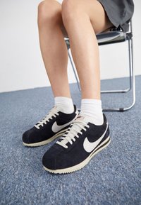 Jalat yang mengenakan kaus kaki putih dan sepatu sneaker hitam Nike duduk di kursi krom dengan lantai karpet biru di latar belakang.