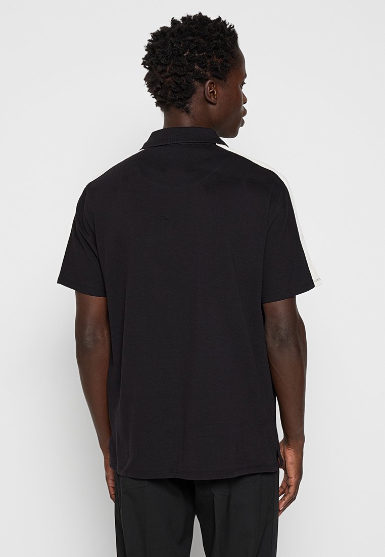 Polo noir à manches courtes avec col, comprenant des accents blancs contrastants sur les épaules et un dos uni. Tissu en mélange de coton.