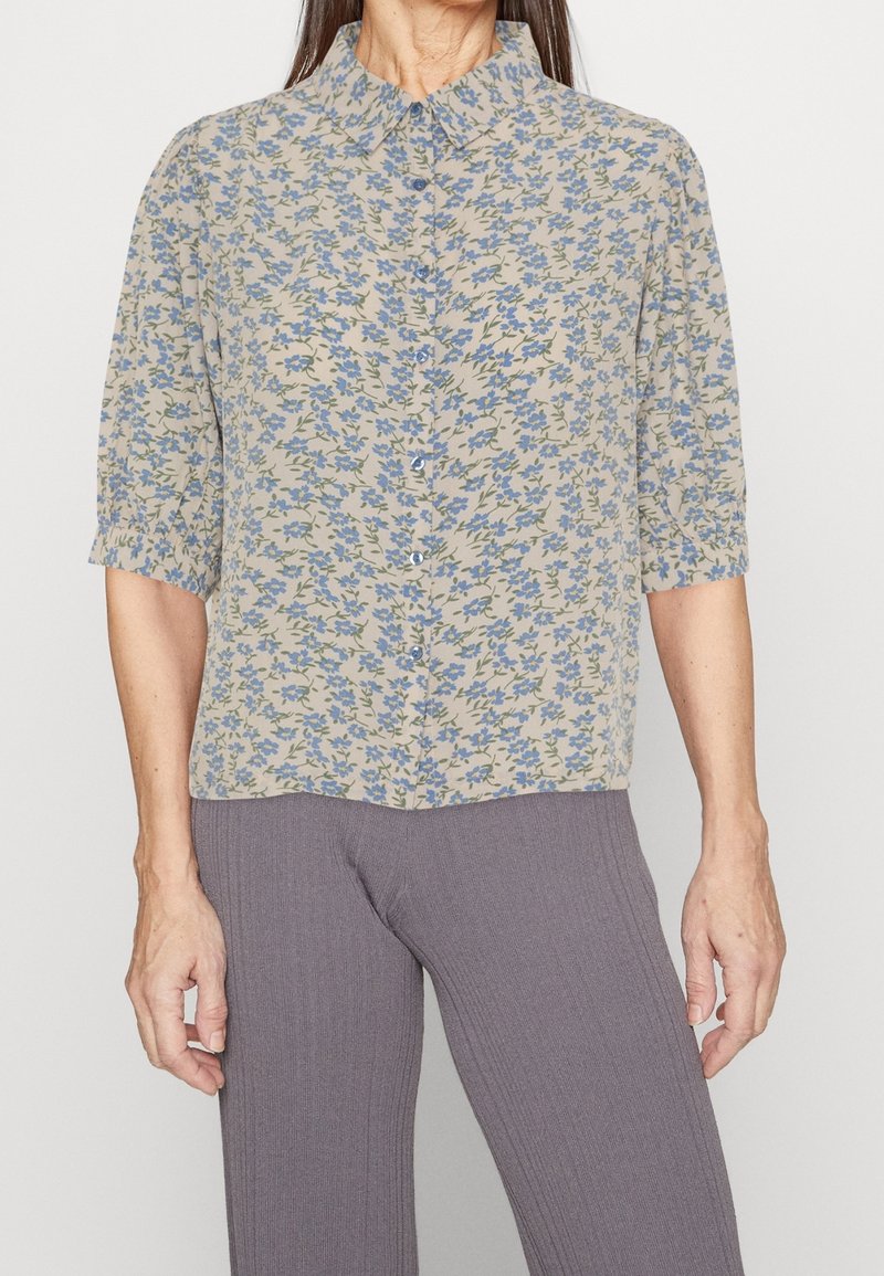 Blouse fleurie avec une base beige clair et un imprimé floral bleu, dotée d'un col à revers, de manches courtes et d'une fermeture boutonnée à l'avant.