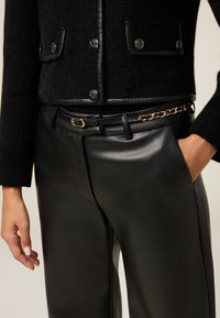 Zwarte textuurjasje met zilveren knopen over een strakke zwarte leren broek. Bevat een gouden kettingaccent op de riem. Glad, strak materiaal.
