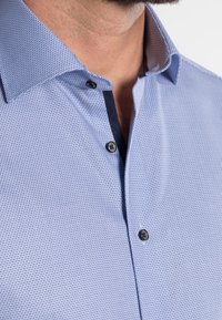 Chemise bleu clair à motif avec un col structuré, deux boutons noirs et des accents foncés le long de la patte de boutonnage, fabriquée dans un tissu texturé.