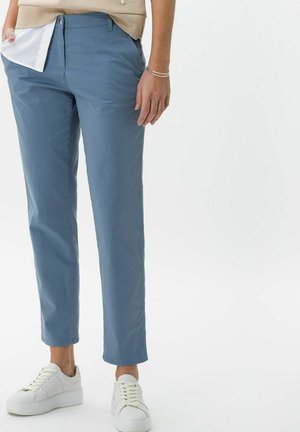 Personne portant un pantalon bleu fuselé, un haut beige, une chemise blanche en dessous et des baskets blanches, debout les mains dans les poches sur fond blanc.