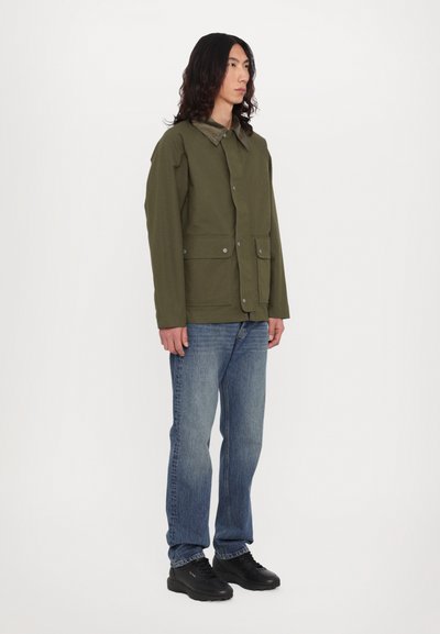 Veste en toile verte avec un col, poches avant et boutons-pression. Portée avec un jean bleu et des baskets noires.