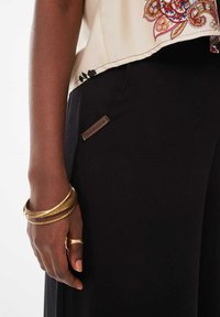 Zwarte broek met een close-up van een hand die meerdere gouden armbanden draagt, met een bruine label met de tekst "blue shadow" aan de zijkant.