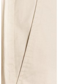 Gros plan sur un tissu beige montrant une couture diagonale avec des surpiqûres détaillées sur une surface textile lisse et tissée serrée.