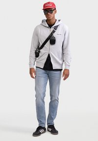 Γκρι φούτερ με φερμουάρ και μαύρο λογότυπο, μαύρο μπλουζάκι, ανοιχτόχρωμα μπλε τζιν, μαύρα αθλητικά παπούτσια με λευκές ρίγες, κόκκινο καπέλο Coca-Cola, μαύρα γυαλιά ηλίου.