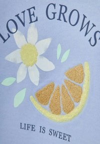Un tessuto azzurro chiaro presenta un design ricamato con un fiore bianco, un centro giallo e una fetta d'arancia. Il testo recita "L'AMORE CRESCE", "LA VITA È DOLCE."