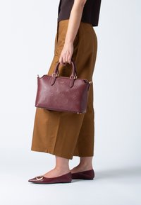 Bordeaux læder håndtaske med en struktureret design, præget mønster, guldfarvet hardware og dobbelte håndtag. Sammen med brune culottes og flade sko.