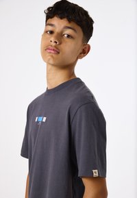 Camiseta de algodón gris oscuro con mangas cortas, escote redondeado y un pequeño gráfico azul y blanco en la parte frontal. Etiqueta en la manga izquierda.