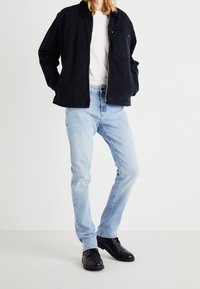 Svart kanvasjacka med en framficka, bärs över en vit t-shirt, i kombination med ljusblå jeans och svarta skor. Enkel, avslappnad outfit.