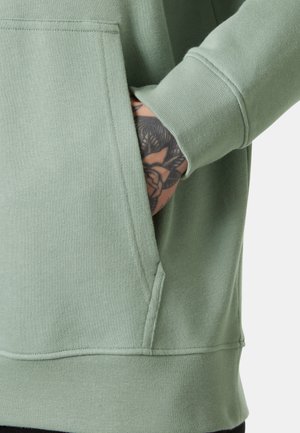 Mano con tatuaje floral negro parcialmente dentro del bolsillo frontal de una sudadera verde clara con puño acanalado.