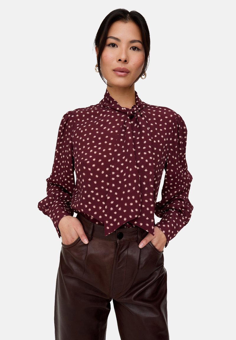 Weinrote Bluse mit weißem Sternmuster, verfügt über einen gebundenen Ausschnitt, lange Ärmel mit gerafften Schultern, kombiniert mit hoch taillierter brauner Lederhose.